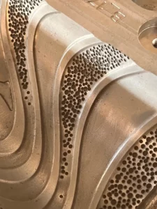 Metalowa powierzchnia złożona z gładkich, zakrzywionych wzorów oraz precyzyjnie rozmieszczonych, okrągłych otworów. Struktura ta jest efektem zaawansowanego teksturowania laserowego.