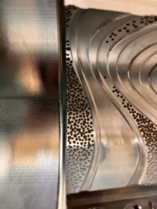 Zbliżenie na metalową powierzchnię z wyraźnym kontrastem między gładkim, lśniącym fragmentem a zakrzywioną częścią z teksturą laserową. Obszar teksturowany zawiera gęsto rozmieszczone otwory w precyzyjnym wzorze, tworząc estetyczny i funkcjonalny efekt.