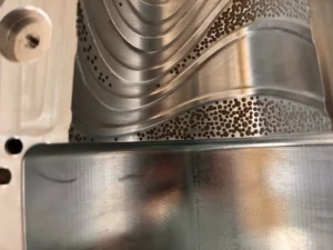 Metalowa powierzchnia z widocznymi wzorami uzyskanymi za pomocą teksturowania laserowego. Struktura obejmuje gładkie krzywizny oraz drobne, precyzyjnie rozmieszczone otwory.