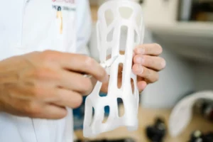 Inżynier trzyma w rękach prototyp wykonany z białego tworzywa w technologii druku 3D, o ażurowej strukturze, prawdopodobnie służący jako medyczna orteza lub element ochronny, i dokonuje precyzyjnych poprawek narzędziem chirurgicznym lub modelarskim.
